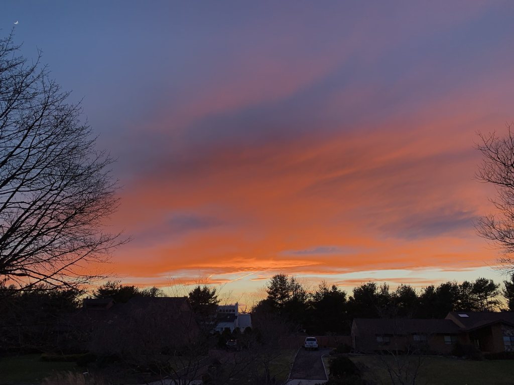 Orange and purple sunset clouds over a quiet neighborhood, with a crescent moon