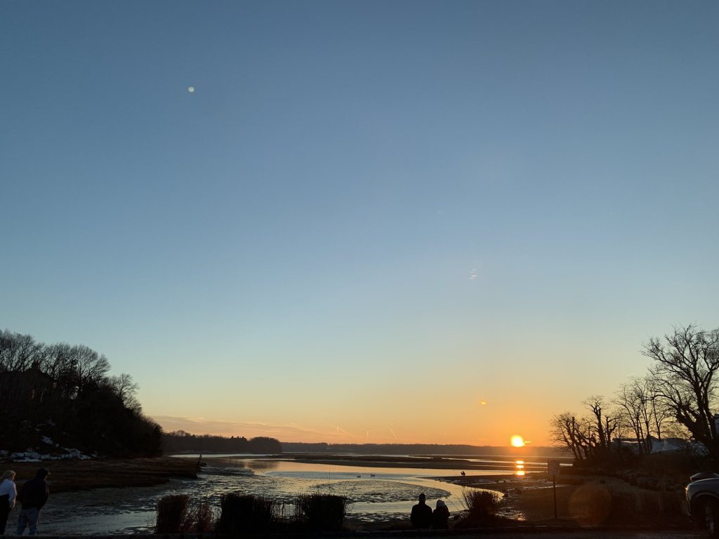 Sun setting over a harbor at low tide, with people watching from a bench