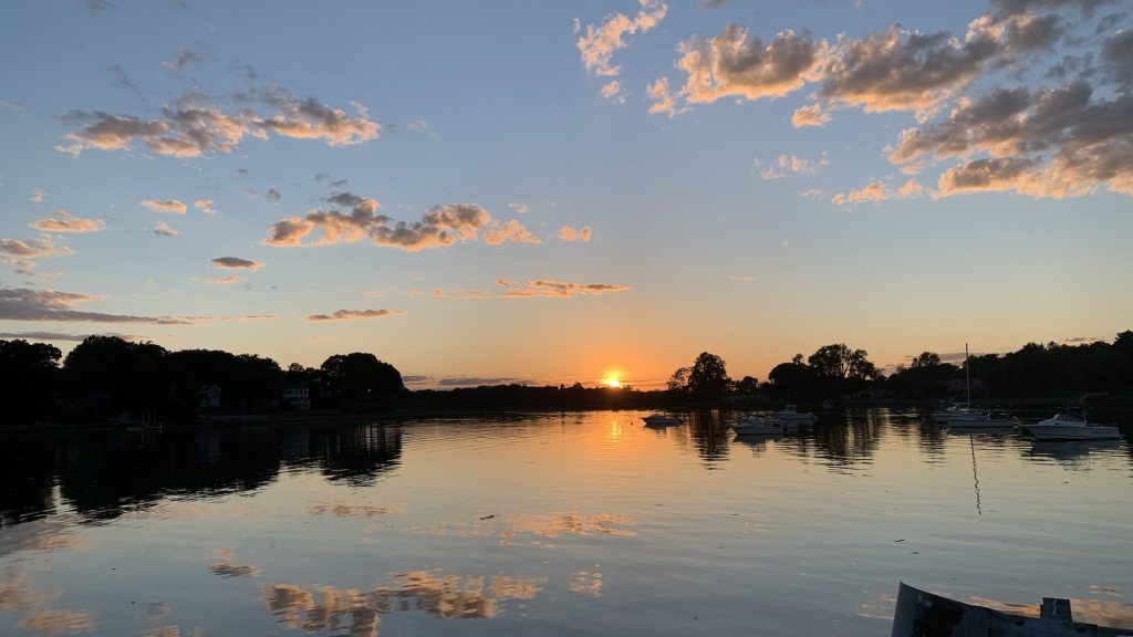 Sun setting over a calm harbor with moored boats and golden clouds